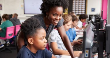 A teacher looking over a student's shoulder as they both look into a computer monitor in a classroom